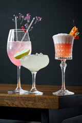 Three cocktails—pink gin and tonic with flowers, a margarita with lime, and a frothy pink drink with cherry and orange—rest on a wooden table and gray background