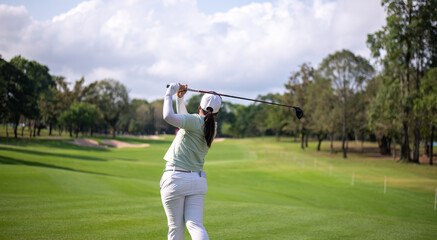 Professional female golfer lush green golf course, mid-swing after hitting a shot through after driving the ball down the fairway trees and distant sand bunkers, typical of a competitive setting.  