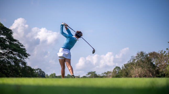 Professional female golfer lush green golf course, mid-swing after hitting a shot through after driving the ball down the fairway trees and distant sand bunkers, typical of a competitive setting.  