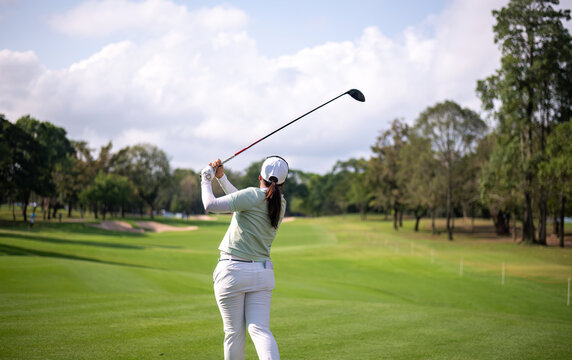 Professional female golfer lush green golf course, mid-swing after hitting a shot through after driving the ball down the fairway trees and distant sand bunkers, typical of a competitive setting.  