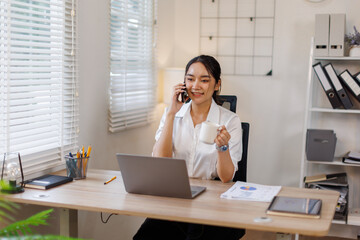 Obraz premium Happy young business woman manager talking consulting client on cell phone working on laptop in the office. Busy woman professional business making call on cell using computer sitting at desk.