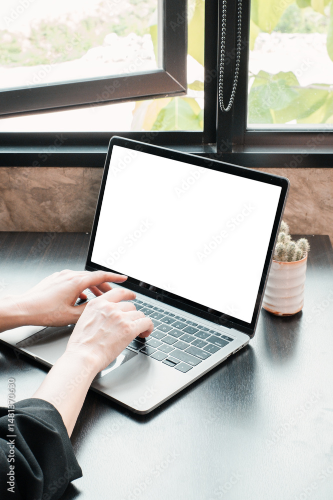 Poster hands typing on a laptop with a blank screen, set by a window in a bright, modern workspace.