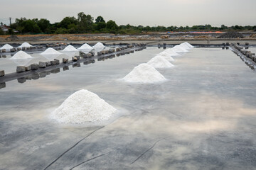 Salt piles grow in shallow pools as seawater evaporates under sun near village. Crystalline salt mounds form in rustic production ponds, mineral deposits from terraced seawater reservoirs, mining