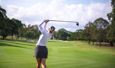 Professional female golfer lush green golf course, mid-swing after hitting a shot through after driving the ball down the fairway trees and distant sand bunkers, typical of a competitive setting.  
