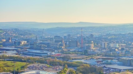 Elevated drone footage showing the Huddersfield Town Football Club area and cityscape, featuring commercial buildings, stadium outlines, cranes, and rolling hills in warm morning light - Powered by Adobe