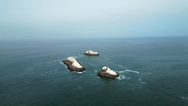 Drone moves forward over calm ocean toward rocky islands with seabirds. Shot in Lima, Peru at 8am under soft, clear morning light.
