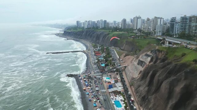 Aerial footage of paragliders soaring over Lima's Costa Verde, with ocean, cliffs, greenery and traffic below on a bright, partly cloudy morning.