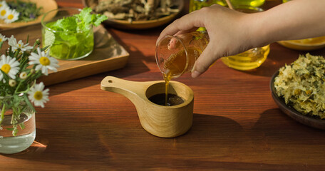 A hand is holding a beaker containing a yellow liquid and pouring it into a wooden cup with a handle. Inside the wooden cup are aromatic particles, mixed to form a brown medicinal mixture.