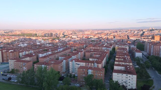 Smooth side‑tracking aerial shot from Cerro del T&iacute;o P&iacute;o: red‑tiled residential blocks of Vallecas glide past in warm evening light while the Madrid skyline glows on the far horizon