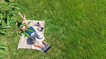 Young woman using laptop computer in park, student girl freelancer working and studying online outdoors sitting on grass with headphones and laptop, aerial drone view from above