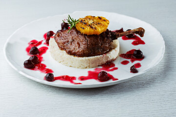 Gourmet duck leg with grilled orange, cherry sauce, and creamy risotto on a white plate, and white wooden table