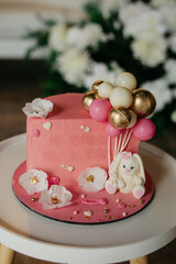 pink cake for celebrating a child's birthday, or a girl's birthday, the cake is decorated with white flowers, a toy bunny, and decorative balloons, the cake is on the table