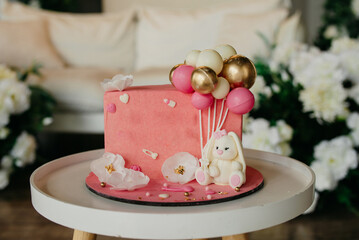 pink cake for a child's birthday celebration, decorated with white flowers, a toy bunny, and decorative balloons, the cake stands on a table against a background decorated with flowers