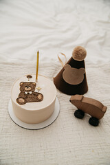 A cake for a child's birthday celebration with one candle and a decorative bear on top standing on an isolated background next to a festive hat on his head and a toy car