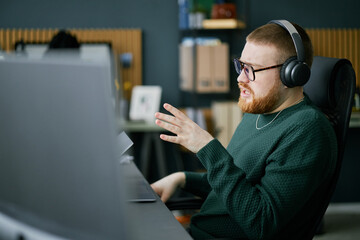 Man wearing glasses and headphones, working on computer at desk in modern office. Bookshelf and wall decor visible in background adding to atmosphere