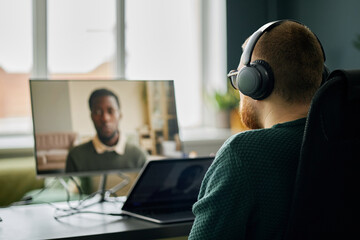 Man wearing headphones sitting at desk with computer and laptop engaged in virtual meeting with...