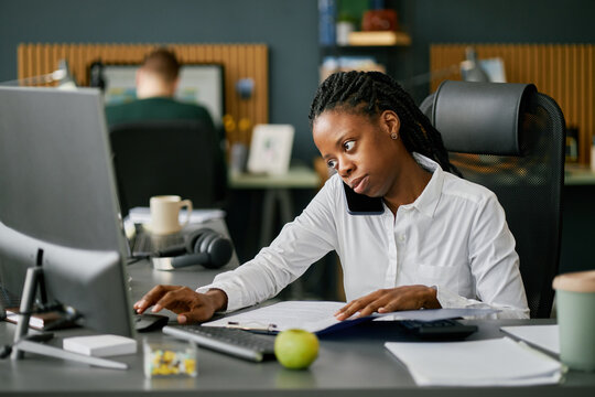 African American woman working in modern office, talking on smartphone and using computer at desk, displaying multitasking and concentration