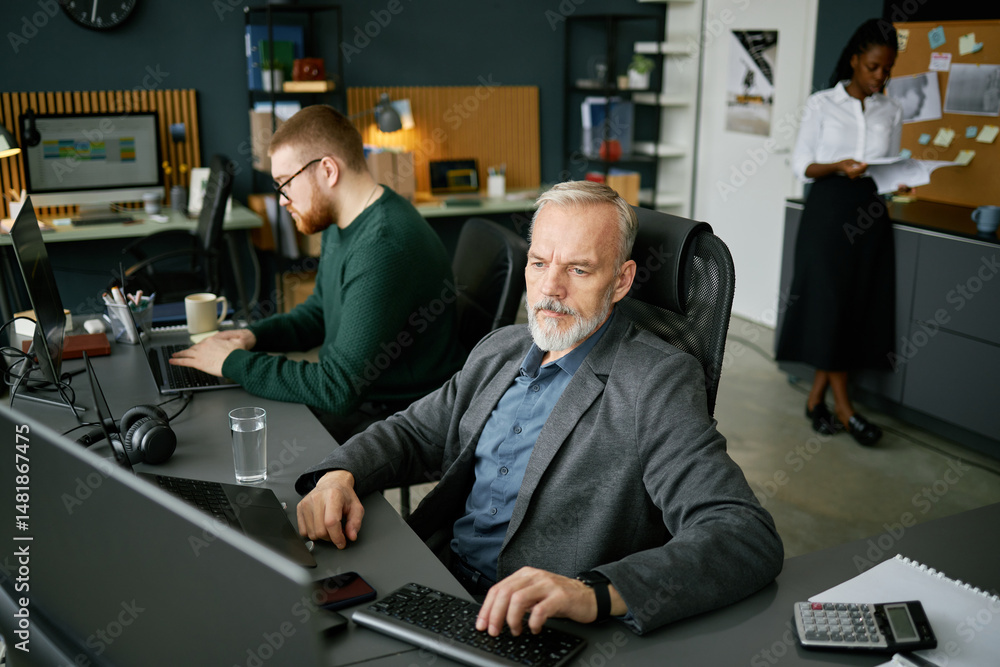 Poster office scene depicting team collaborating on projects with focus on mature man at desk along with co