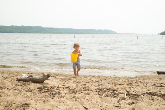 Young boy carries yellow bucket along a quiet lakeshore beach.