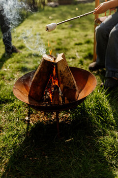 Person roasting marshmallow over a backyard fire pit in sunlight