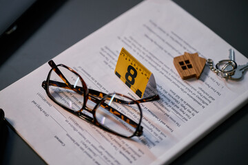 Close up of a pair of glasses and an evidence marker, placed on an open book near a keychain with small house shaped pendant and other items on dark surface
