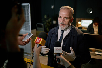 Elderly businessman standing near reporters, holding interviews in an office environment. Discussing important matters while multiple reporters holding microphones and recording