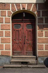 Worn red wooden double doors framed by a textured archway glow in warm sunlight. Cracks, scratches, and old stone steps whisper stories of age, mystery, and quiet European charm.