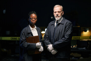 Multiracial detectives standing side by side, holding clipboard and wearing gloves in dimly lit...