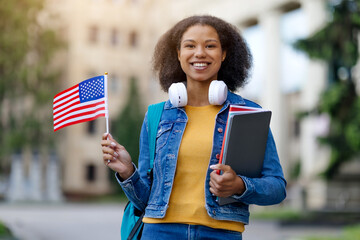 Study Abroad. Smiling Black Female Student Posing With American Flag Outdoors, Happy Young African American Woman With Backpack And Workbooks Standing Near University Building, Copy Space