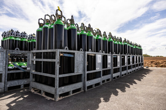 Numerous green and white topped black gas cylinders are secured in metal crates and stored outdoors on a paved surface under a partly cloudy sky