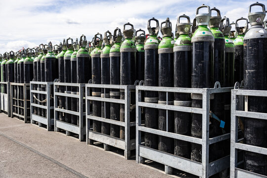 Numerous green and white topped black gas cylinders are secured in metal crates and stored outdoors on a paved surface under a partly cloudy sky