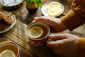 Drinking tea during morning breakfast in rustic cozy kitchen. Female hands holding mug of herbal tea with lemon slice