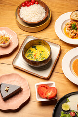 Top view of Sri Lankan coconut curry with rice, chocolate cake slice, prawns chimichurri, and raw tomato in a bowl, all placed on a brown wooden table in a restaurant.