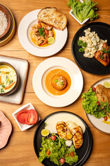 Top view of Sri Lankan coconut curry with rice, arancini, prawns chimichurri, pan-seared fish, white cheese macaroni with chicken, raw tomato, chocolate cake on a brown tab