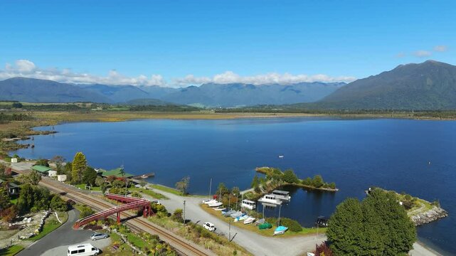 Smooth forward drone shot glides over the calm waters of Lake Brunner in New Zealand, revealing distant views of the snow-capped Southern Alps under clear skies in a tranquil natural setting.