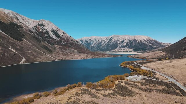Aerial left orbit establishing shot of Lake Pearson in Arthur&rsquo;s Pass, New Zealand, showcasing golden autumn colors, mountainous terrain, and the calm alpine lake under clear skies.