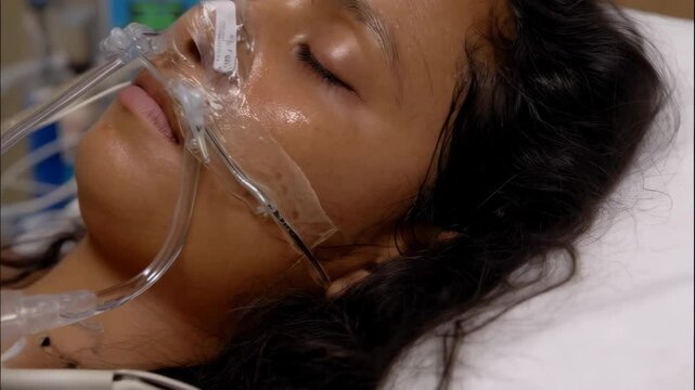 Ill woman with dark hair receiving respiratory support in hospital bed, with medical equipment and sensors visible, closed eyes.