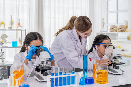 young female teacher helps kid to conduct chemical experiment with microscope,primary school classroom and children in lab coats studying science