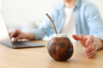 Man using laptop at wooden table indoors, focus on traditional yerba mate tea
