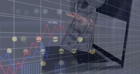 Showing adult hand inserting US dollar bills into open laptop on tabletop, with line charts overlay