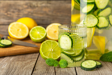 Cucumber water with lemon, lime and mint on a wooden table. Water infused with sliced ​​cucumber in a drinking glass. Tonic refreshing drink. Cucumber lemonade.Summer natural cooling drink.Copy space