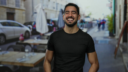 Fototapeta premium Young man smiling outdoors on a restaurant terrace, showcasing casual style and vibrant energy in an urban setting with blurred city background.