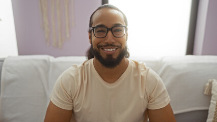 Young man with beard smiling indoors in a cozy living room wearing glasses and a casual shirt