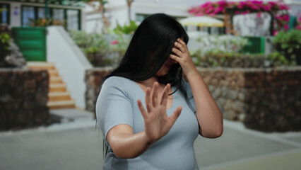 Woman covering face with hand outdoors, appearing scared, standing on city street, dressed...