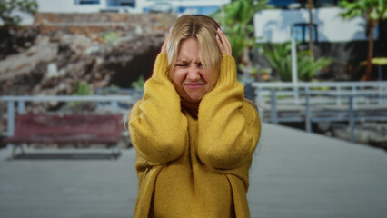 Woman holding her head in frustration on a city street wearing a yellow sweater with trees and...