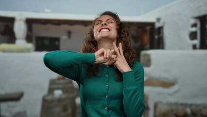 Woman smiling showing hashtag gesture outdoors wearing green sweater in urban street setting with white background reflecting city life and vibrant expression.