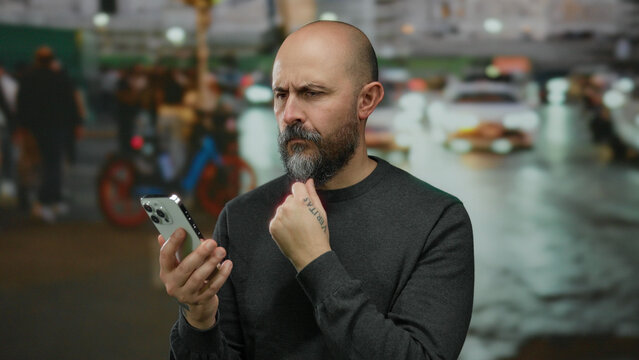 Bald man with beard intently looking at smartphone on city street at night, surrounded by urban activity and bright lights, embodying a thoughtful demeanor outdoors.