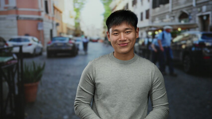 Fototapeta premium Young man smiling outdoors on a bustling city street, reflecting a cheerful and serene mood amidst urban backdrop with cars and buildings, wearing a grey sweater in daytime.