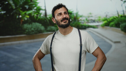 Fototapeta premium Hispanic man with beard wearing suspenders smiles outdoors on urban street with lush greenery in background under daylight