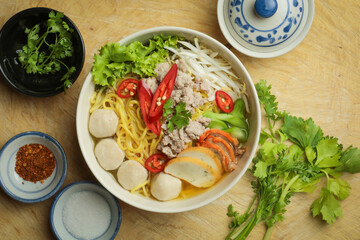 Delicious bowl of Asian noodle soup with fish balls, minced pork, fresh vegetables, and spicy red chili, served on a rustic wooden table with chopsticks.